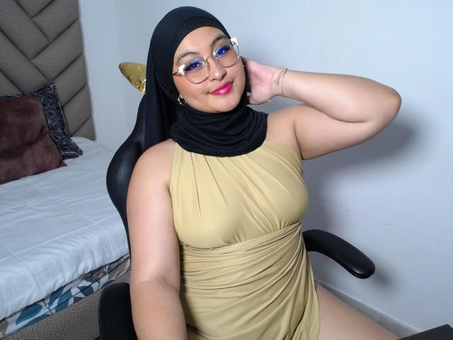 HelueJamil with black hijab, glasses, blue eyeshadow, and gold dress, sitting in chair, hand on neck, smiling, showing her curvy body and small tits, white wall background.