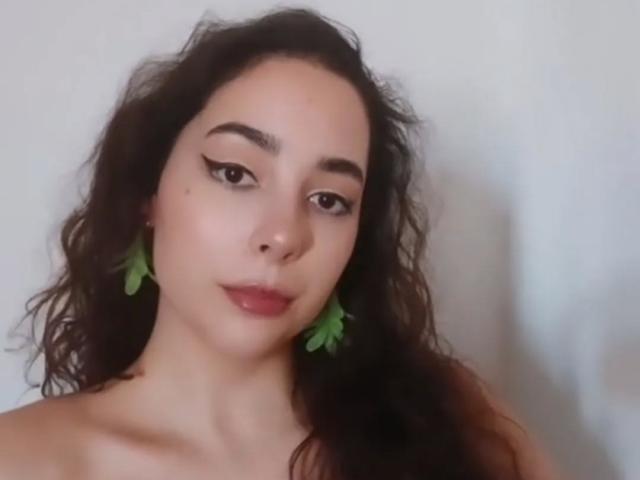 SyrenaValentina with wavy dark hair, green leaf earrings, and winged eyeliner, showing off her glossy red lips and smooth, light-skinned face. She's looking slightly to the side.