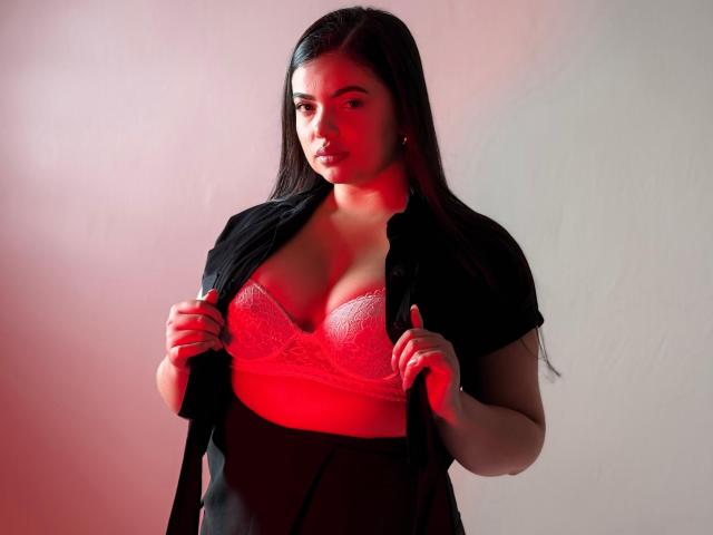 LaraJames with long dark hair, wearing a black shirt open to reveal pink lace boobs, holding the shirt with both hands, standing under red and white lighting.