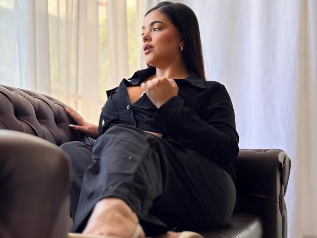 Lara James, con cabello negro largo, sentada en un sofá de cuero marrón, vestida con una camisa negra abotonada y pantalones negros, sosteniendo su camisa, mirando hacia un lado, mostrando su trasero..