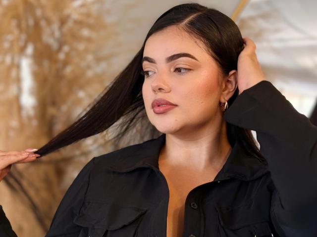 Lara James, com longos cabelos pretos, vestindo uma camisa preta, segurando o cabelo com uma mão, tem batom rosa, brincos de ouro e uma camisa ligeiramente aberta com alguns botões desfeitos..