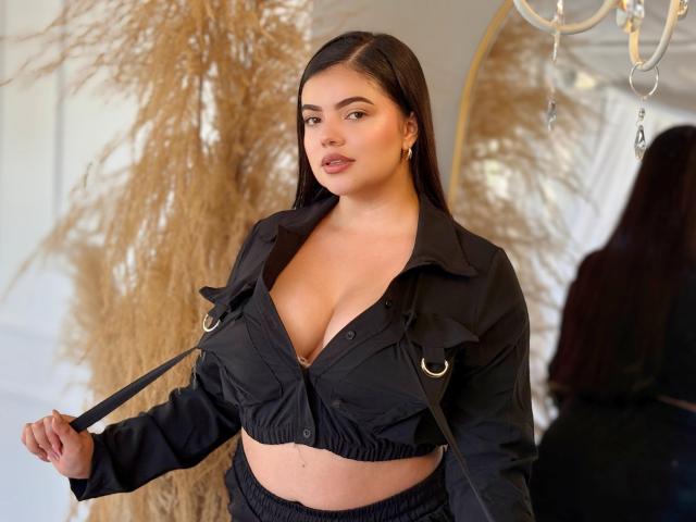 LaraJames with dark hair, wearing a black crop top showing her big boobs, holding the straps, standing in front of a mirror, looking confident.