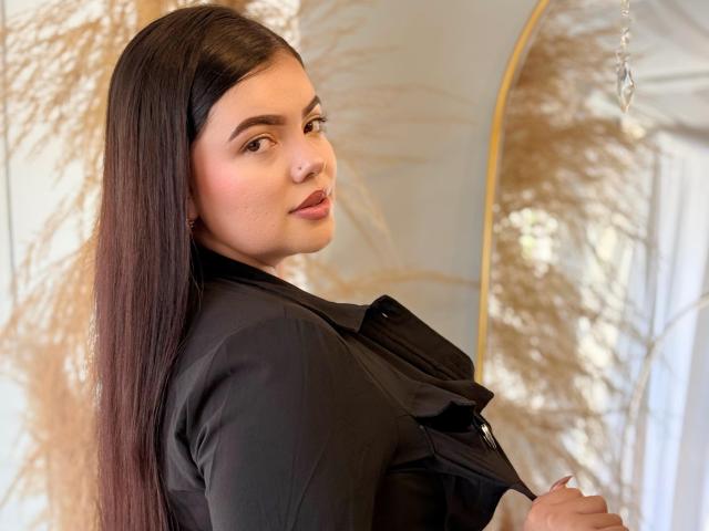 LaraJames with long dark hair, black shirt, looking over shoulder, slightly smiling, holding shirt, subtle makeup, cute, small nose ring, puffy ass, soft light, relaxed, attractive.