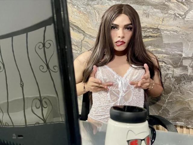 SahraValley with long brown hair, wearing white lace lingerie, touching her tits with fingers pointing upwards, sitting in front of a table with a white mug.