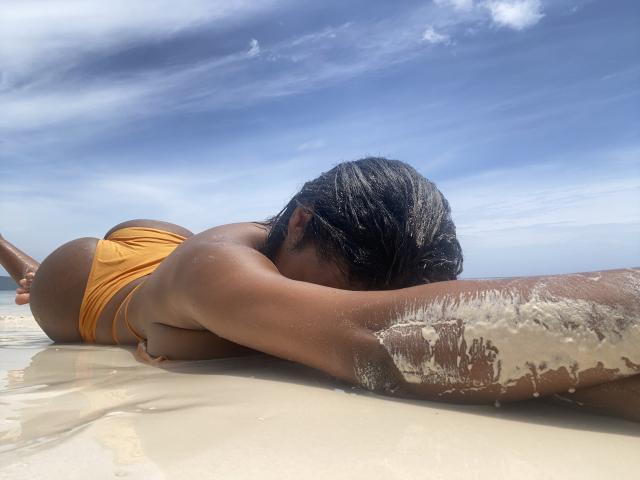 NahomyCruz lying face-down on a sandy beach, wearing an orange bikini, with wet sand on her arm, and her ass prominently raised, under a blue sky.