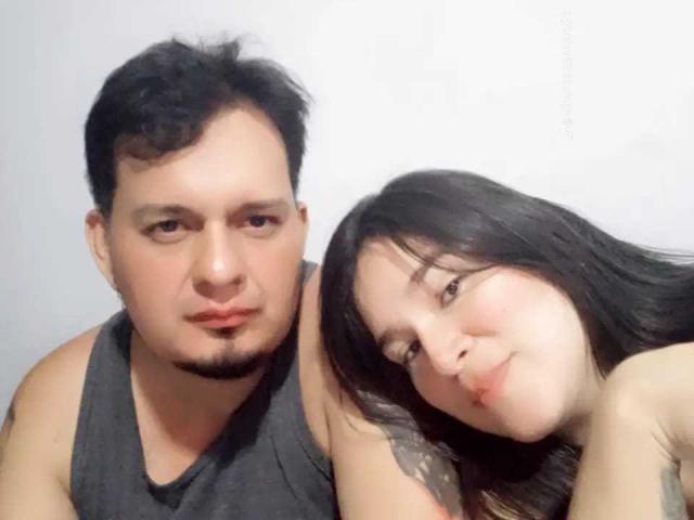 SofiayJimmy has a chubby face, black hair, and a goatee, wearing a grey tank top. SofiayJimmy's wife, with black hair and tattoos, leans on him, smiling.