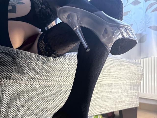 Las medias de red negras hasta el muslo de MadameSolenne1-hot, con tacón alto transparente y tirantes de encaje, mostrando su muslo tatuado y ropa interior negra, sentada en un sofá gris con textura..