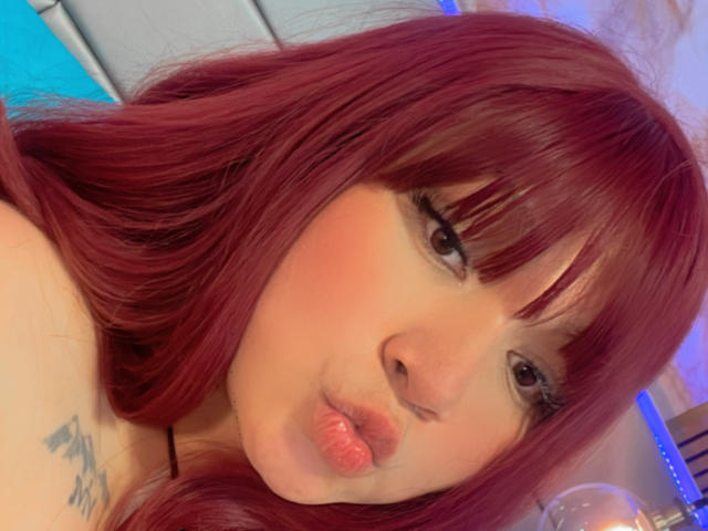 LillyMayer with red hair, pouty lips, tattoo on shoulder, looking at camera, soft pink blush on cheeks, eyeliner, black necklace, blue light in background.