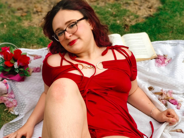 AlanisSweet with red hair, glasses, and red dress, sitting on a blanket, leg raised, showing her tits, small book, flowers, grass, and white blanket in the background.
