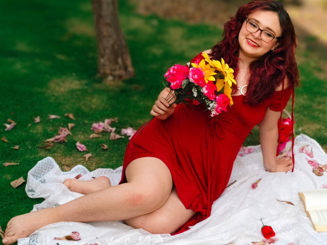 AlanisSweet with red dress, glasses, and curly red hair, holding flower bouquet, sitting on white lace blanket, legs crossed, book to the right, grassy background.