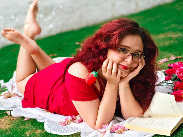AlanisSweet, red-haired, lying on grass, wearing red dress, glasses, hands on face, book, and strawberry on shoulder, legs up, white blanket, pink flowers, bare feet, cute, sexy.