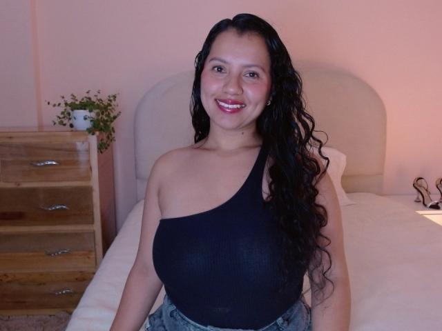 Isis69s with long black wavy hair, smiling, wearing a black one-shoulder top, showing her big tits, sitting on a bed, wooden dresser with plant on left.