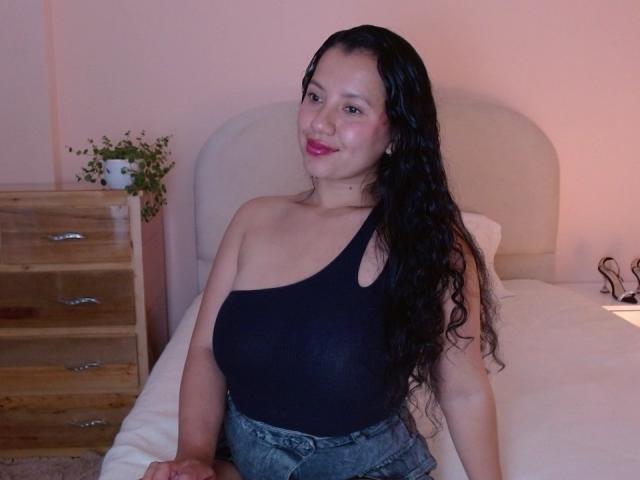 Isis69s with long black hair, wearing a black one-shoulder top, showing her big tits, and blue jeans, sitting on a bed, smiling with pink lipstick.