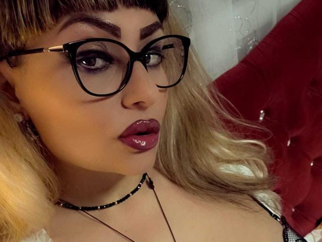 MirayDesire with glossy dark red lipstick, black choker, and black-framed glasses, looking seductively at the camera, showing off her smooth skin and blonde hair with dark roots.