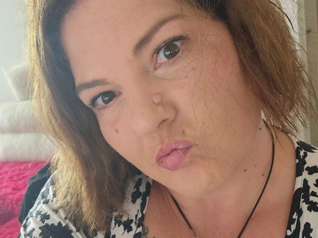 El rostro de Joyfull-hot, con cabello castaño y ondulado, piel clara y lápiz labial rosa, muestra una expresión de labios fruncidos, como si estuviera besando. Lleva una blusa con estampado blanco y negro y un collar..