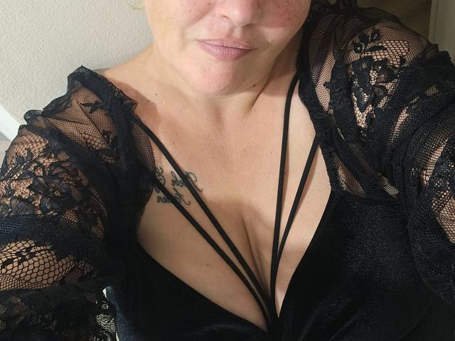 Joyfull-belle, peau claire, petit tatouage sur la poitrine gauche, portant un haut en dentelle noire avec un décolleté plongeant, un haut noir à bretelles, lèvres légèrement entrouvertes, clavicules visibles et partie supérieure de la poitrine..