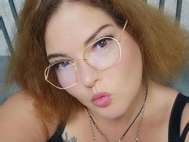 Vrolijk uitziend meisje met krullend bruin haar, gouden bril, roze lippenstift, volle lippen, een zwart tanktopje, een ketting en een tatoeage op de linker schouder, die omhoog kijkt met lichtjes open mond..