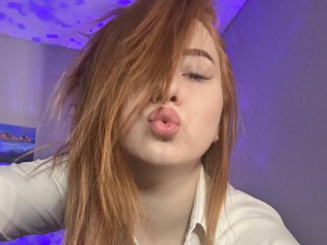 Barberry with red hair, pouty pink lips, and long eyelashes, making a kissy face, wearing a white shirt, in a purple-lit room. Her hair is partially covering her eyes.