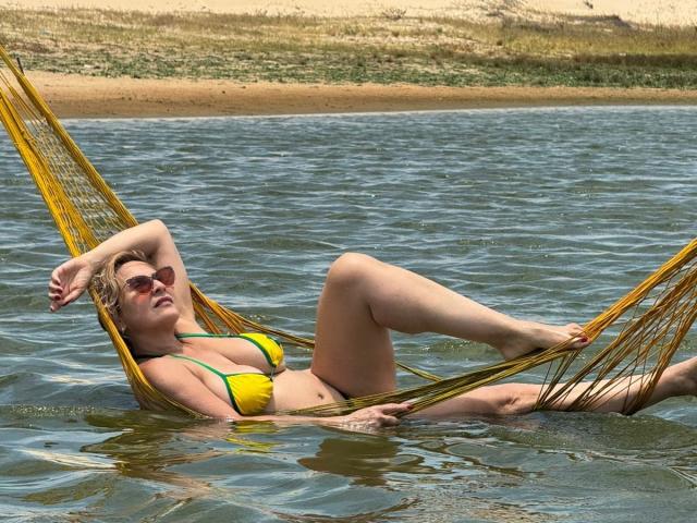 JaneTurner in a yellow bikini, lying in a yellow hammock, water up to her ass, legs crossed, arm behind head, wearing sunglasses, blonde hair, relaxed, sunny.