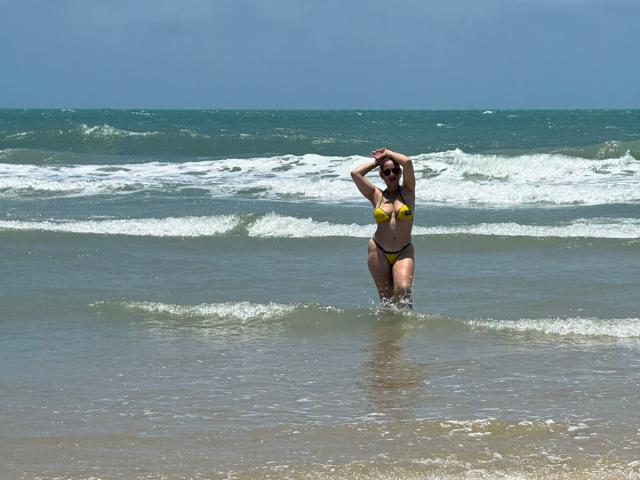 JaneTurner in a yellow bikini with yellow top and black bottom, standing in shallow ocean water, arms raised, showing her tits and ass, with waves in the background.
