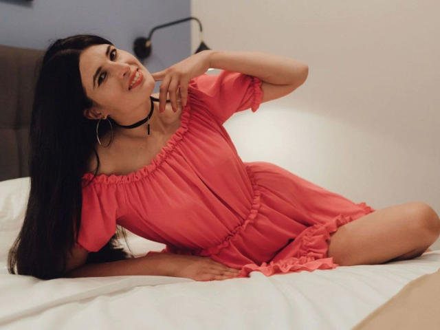 DarkWhiskers-hot with long black hair, pink dress, and black choker, sitting on a bed, smiling, showing her tits, and touching her neck with a hand.