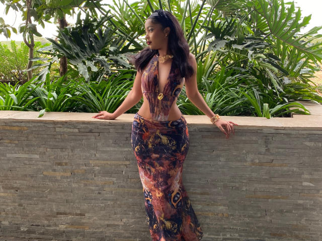 PreatyLexxi with long dark hair, wearing a deep V-neck, colorful, sheer dress, and gold jewelry, leaning against a stone wall, looking to the side, hands on wall.