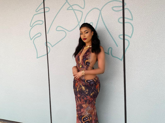 PreatyLexxi stands in a colorful, patterned dress, showing her ass and tits, with long black hair, red lipstick, and gold jewelry, against a white wall with green graffiti.