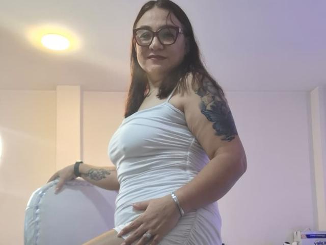 LadyNylonOh, usando óculos, vestindo um vestido branco, mostrando os seios, segurando um travesseiro branco com babados, tatuagem no braço, pulseira no pulso, em pé em um quarto, olhando para a câmera..
