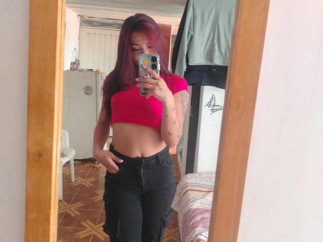 SashaHans with pink top, black pants, showing a flat stomach and tattoos, taking a mirror selfie, holding a phone with stickers, pink hair, white room, wooden mirror.