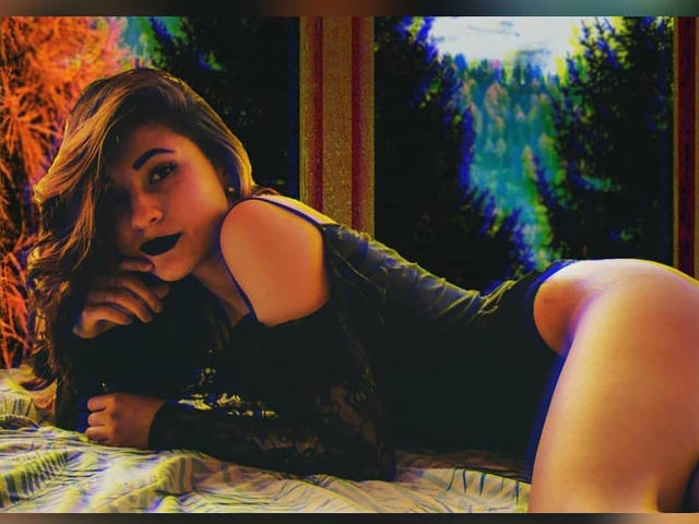 LilithStorm lying on bed, wearing black lace top, exposing her ass, with dark lipstick, long brown hair, looking at camera, forest outside window, colorful light reflections.