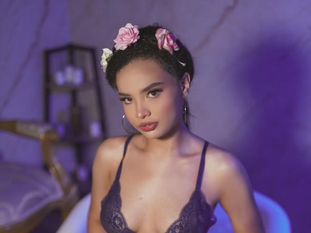 DulceNayra with curly black hair, pink and white flower headpiece, wearing a purple lace bra, looking seductively at the camera, small tits, hoop earrings, purple lighting.