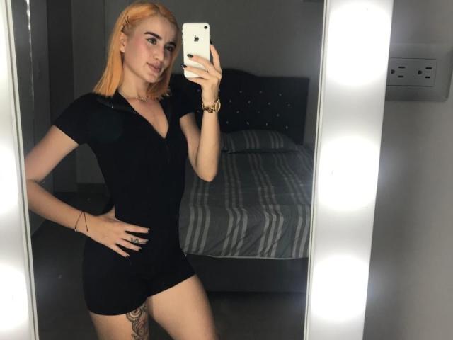 AnitaLux with orange hair, black dress, and gold bracelet, taking a mirror selfie, showing a tattoo on her thigh, hand on hip, holding a white iPhone.