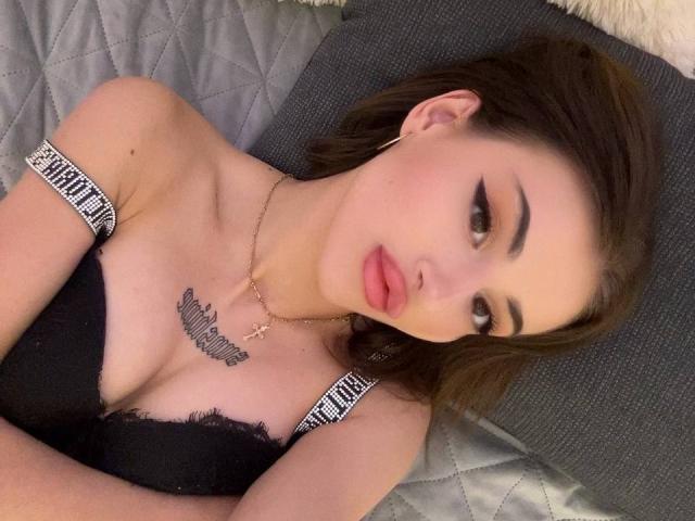 SunshineLi with brown hair, black bra, and pink lipstick, lying on a gray pillow, wearing a cross necklace, and a tattoo under her tits, looking at the camera.