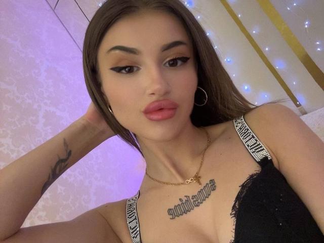 SunshineLi with brown hair, pink lipstick, black lace bra, and a tattooed chest, wearing a gold necklace and hoop earrings, looking at the camera with a pouty expression.