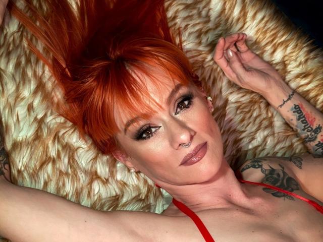 ScarlettN with bright red hair, lying on a fluffy white pillow, showing tattoos, nose ring, and red lipstick, wearing a red top, arms raised, looking at the camera.