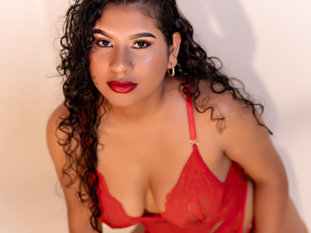 RenataPalmer with curly black hair, red lipstick, and red lace bra, showing her boobs, looking up with a slight smile, gold hoop earrings, warm light.