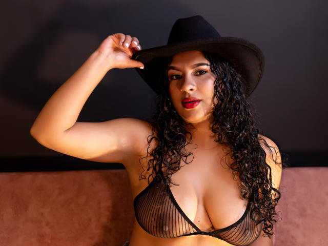 RenataPalmer with curly black hair, wearing a black hat, red lipstick, and a sheer black bra, showing her big tits and a confident, sexy expression.