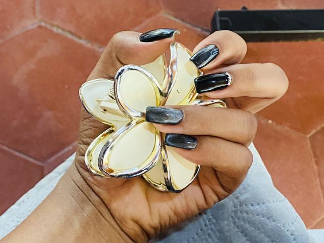 La mano de NatalyBlonder, con uñas negras brillantes, sostiene un objeto amarillo y plateado con forma de flor, y su piel bronceada contrasta con las superficies brillantes y reflectantes..