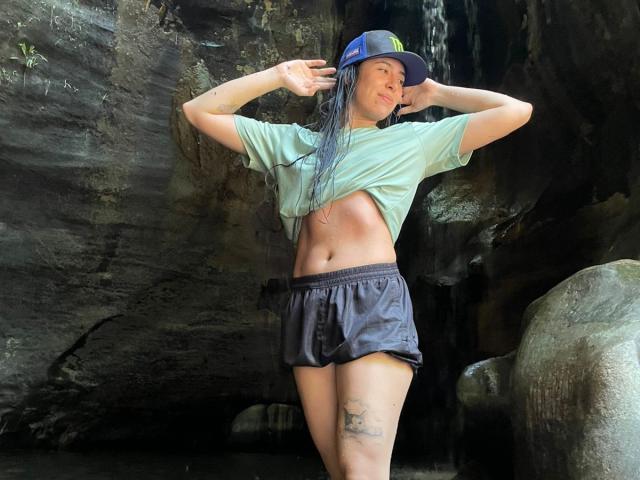 MiluSky mit nassen, langen, schwarzen Haaren, einem grünen Hemd, das angehoben ist, um definierte Bauchmuskeln zu zeigen, schwarzen Shorts und einer blauen Mütze, steht in einer felsigen Höhle mit einem kleinen Wasserfall..