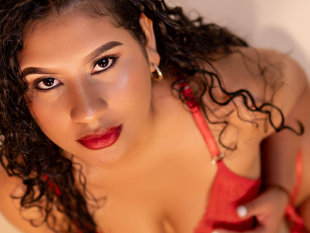 RenataPalmer with curly black hair, red lipstick, and red lingerie, looking up with dark eyes, gold earrings, and red bra straps visible, holding her tits gently.