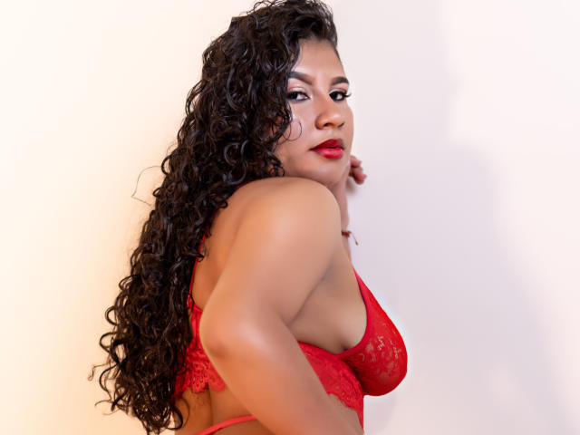 RenataPalmer with curly black hair, red lace bra, and red lipstick, looking over her shoulder, showing her boobs and a slight view of her ass.