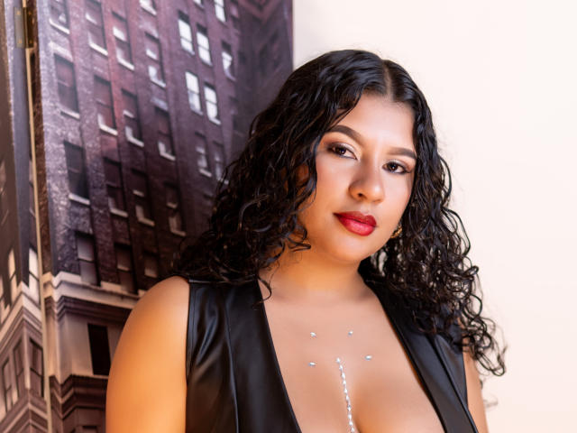 RenataPalmer with curly black hair, red lipstick, and black leather dress, showing her tits with sparkling body paint. She's looking at the camera with a slight smile.