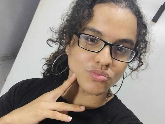 Diane69, met krullend zwart haar, draagt een zwarte bril en een zwart shirt, ze frumelt met haar roze lippen en raakt haar nek aan met een vinger, en draagt grote oorringen..