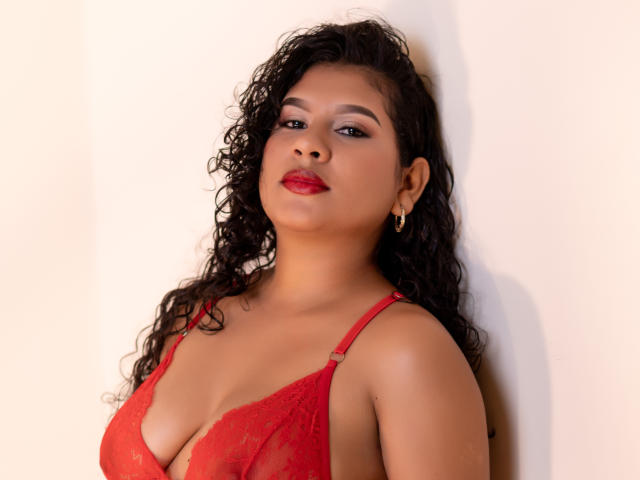 RenataPalmer with curly black hair, red lace bra, and red lipstick, standing against a wall, looking up with a slight smile, wearing gold earrings, showcasing her boobs.
