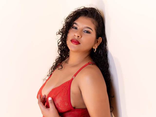 RenataPalmer with curly black hair, red lace bra, and red lipstick, holding her tits against a white wall, wearing gold earrings, looking at the camera with a slight smile.