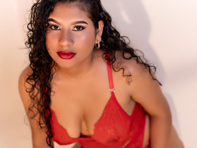 Renata Palmer, avec ses cheveux noirs bouclés, son soutien-gorge en dentelle rouge et son rouge à lèvres rouge, regarde vers le haut avec un léger sourire, mettant en valeur sa poitrine et une boucle d'oreille en or..
