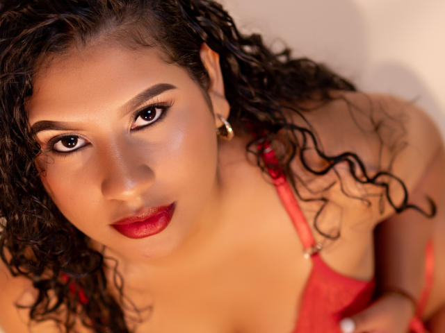 Renata Palmer mit lockigem, schwarzem Haar, rotem Lippenstift und einem roten BH, die mit dunklen, geschwungenen Augenbrauen und goldenen Ohrringen nach oben schaut, wobei ihre Brüste zu sehen sind und sie ein subtiles, verführerisches Lächeln zeigt..
