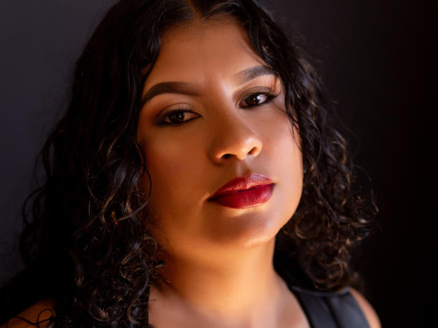 Renata Palmer tem cabelos pretos e cacheados, olhos castanhos e batom vermelho. Seu rosto está iluminado por uma luz suave, que realça sua pele lisa e bronzeada e seus lábios cheios. Ela olha diretamente para a câmera..