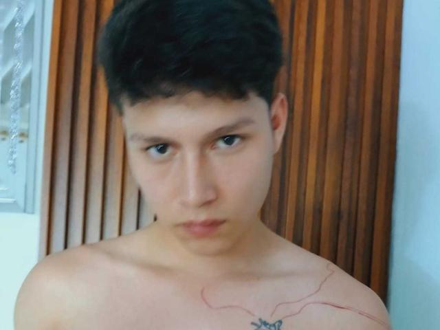 Daniel, sin camisa, con cabello corto y oscuro, expresión seria, piel clara, un tatuaje pequeño en el hombro derecho y una cicatriz visible en la mejilla izquierda, de pie frente a listones de madera..