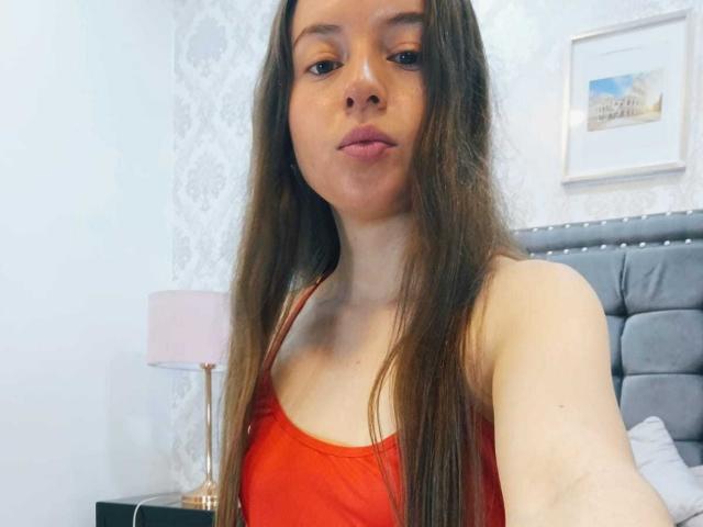 VioletaNova with long brown hair, wearing a red tank top, showing her small tits, pouty lips, and light skin, standing in a bedroom with a pink lamp.